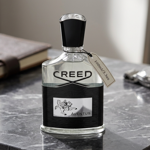 A bottle of Creed Aventus perfume stands on a dark marble surface, with a leather notebook and a silver watch blurred in the background.