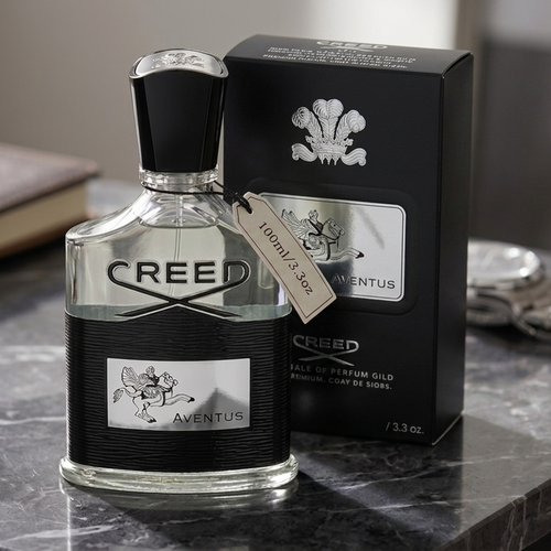 A bottle of Creed Aventus Eau de Parfum with its black retail box, displayed on a dark marble surface next to a notebook and a silver watch.